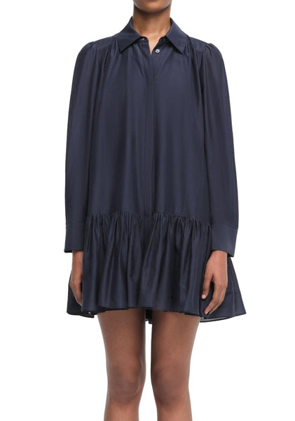 Person wearing a long-sleeve, navy-blue Simkhai Viola Dress with a loose fit and ruffled hem. The dress features a pointed collar and button-down front. The person's face is not visible.