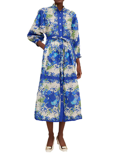 A person is wearing the Cara Cara Beatrice Dress, a classic long blue floral print shirtdress by Cara Cara. It features a button-down front and a tied waist, with subtle ruffle details on the long sleeves. The dress reaches mid-calf and is paired with white shoes.