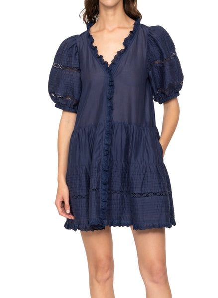 A woman wearing the Sea NY Antonina Dress featuring a navy blue color, short puff sleeves, ruffled hem, lace accents, and a V-neckline with a tiered mini skirt.