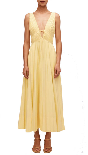 Person in a sleeveless, pleated yellow Simkhai Stephanie Dress with a deep V-neckline and tie detail at the waist, featuring a modern A-line silhouette, standing against a plain white background.