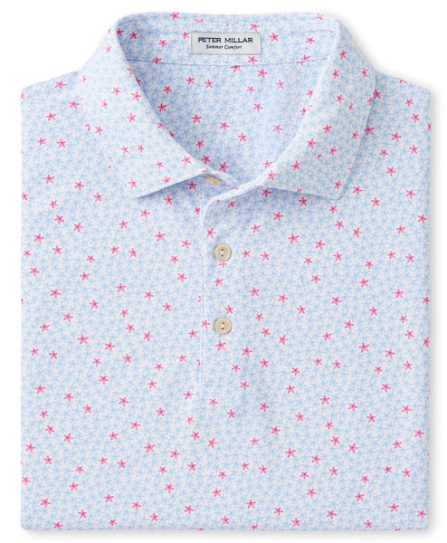 The Peter Millar Starfish Performance Polo in blue is folded and features a pattern of small red stars and tiny light blue floral designs. It includes a three-button placket, four-way stretch for added comfort, and has a label inside the collar reading “Peter Millar Summer Comfort.”