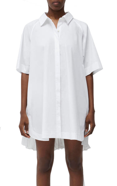 A person wearing the Simkhai Blanche Shirt Dress, a loose-fitting, short-sleeved white button-down shirt dress that embodies casual elegance with its relaxed oversized fit.