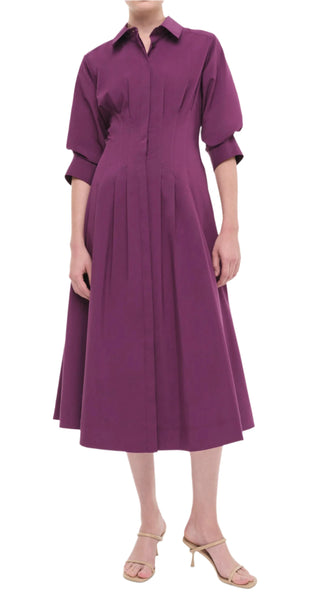 A person wearing the Simkhai Signature Jazz Dress, a purple, knee-length midi dress with elbow-length sleeves and elegant pintuck detailing, paired with beige heeled sandals from Simkhai.