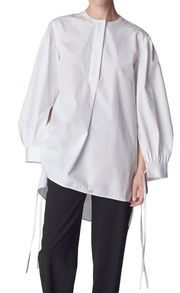 A person is wearing the CO Cuff Tie Shirt, a white, long-sleeve tunic blouse with a front pleat detail, paired with black pants.