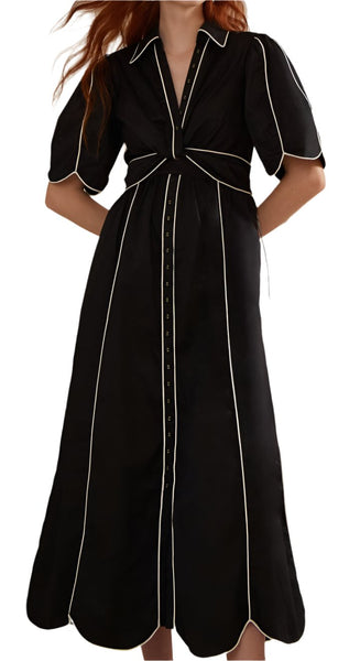 A person is wearing the Farm Rio Black and White Short Sleeve Midi Dress by Farm Rio, featuring white piping, short draped sleeves, a collar neckline, and button details along the front. The dress has a cinched waist and flairs into a midi-length skirt.