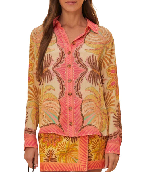 A woman wearing a pink and yellow button-up Farm Rio Palm Scarf Sand Shirt with a multi-colored, intricate embroidered pattern stands facing forward. A matching skirt with similar patterns completes the outfit, capturing the essence of Brazilian fashion in a tropical paradise.