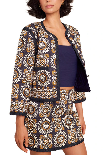 A person wearing a patterned Cara Cara Marissa Jacket and matching skirt set with a navy blue top. The outfit features intricate, circular designs in shades of yellow and white, accented with quilted cotton details.