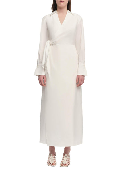 A woman wearing a long-sleeved, white Simkhai Briar Dress with a V-neckline and wrap-style front stands against a plain white background. She is also wearing white sandals.