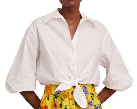 A person wearing a white **Cara Cara Rumson Top** with self-covered buttons, tied at the waist, and a yellow floral skirt. The person's hands are on their hips. The face is not visible.