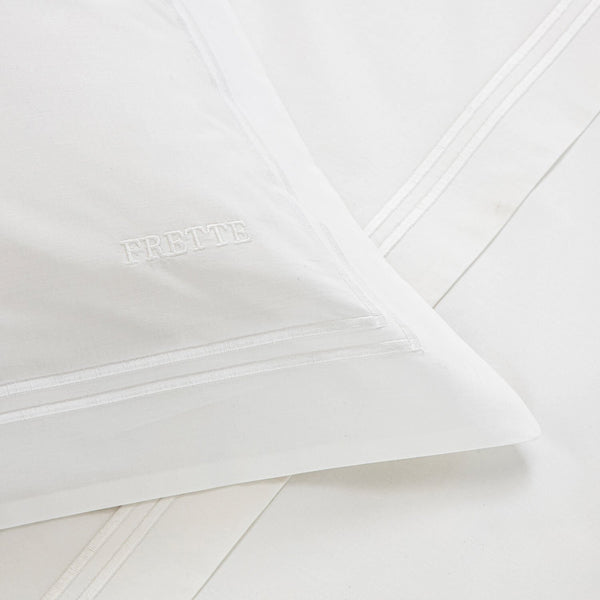 Close-up of a white pillowcase from the Frette Classic Bedding Collection, White/White with embroidered text "FRETTE" on it, lying on matching white bedsheets made from 100% cotton with subtle striped detailing.