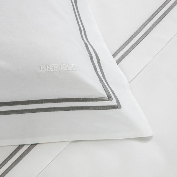 White pillow with double gray border lines, featuring "FRETTE" embroidered in white. Part of the Frette Classic Bedding Collection, White/Grey from Frette, this luxurious sheet set is crafted from 100% cotton for a sumptuous touch.