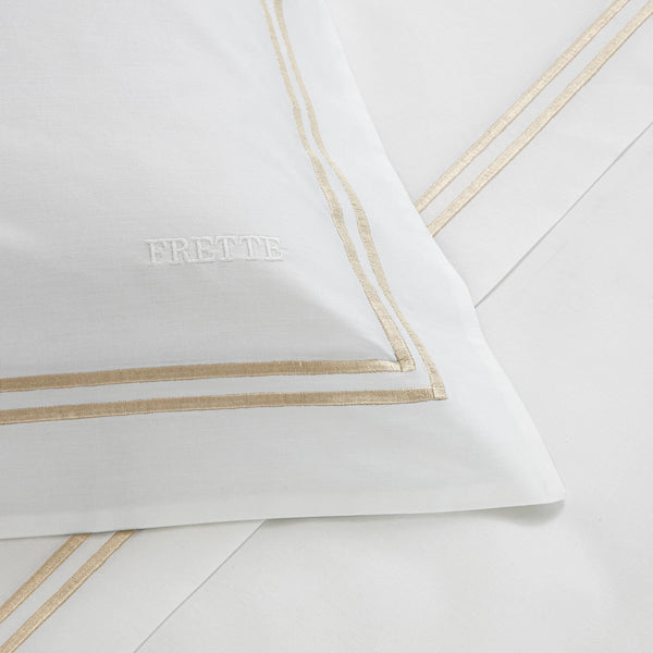 Close-up of a white pillowcase with beige embroidered lines from the Frette Classic Bedding Collection, White/Khaki, featuring the brand name "Frette" stitched on it, placed on matching luxurious 100% cotton sheets.