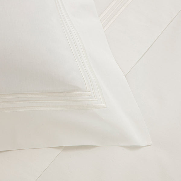 Close-up of a white pillowcase with subtle stitched lines along the edges, laying on a matching white sheet from the Frette Triplo Bourdon Bedding Collection, White/Milk. This elegant piece embodies the luxury of Italian-made bedding, crafted from delicate cotton poplin sheets by Frette.