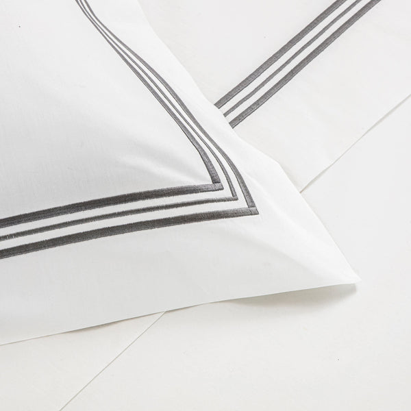 Close-up of a white bed sheet and pillowcase from the Frette Triplo Bourdon Bedding Collection, White/Grey, featuring simple black three-line embroidery along the edges.