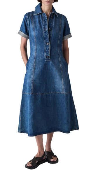 A person wearing the CO Denim Midi Dress by CO, featuring short sleeves, a collared button-front design, and hands in the pockets, paired with black sandals. The dress is made from 100% cotton.