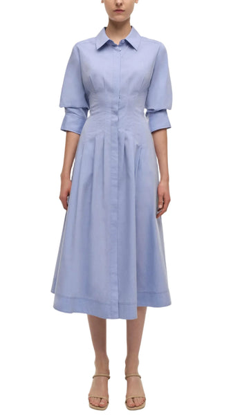 A woman models a light blue Simkhai Signature Jazz Dress by Simkhai, featuring a classic collar and button-down, long-sleeve design. The tailored bodice complements the pleated skirt, while her hands rest by her sides and she wears beige, open-toe sandals.