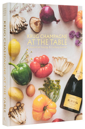 The cookbook "Krug Champagne at the Table" by Rizzoli is beautifully showcased among a colorful assortment of vegetables and a bottle of Krug Grande Cuvée, providing readers with a culinary journey around the world through the expertise of master chefs.