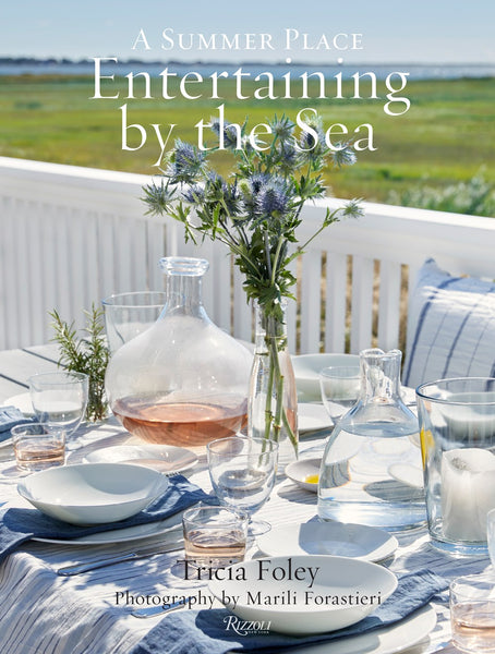 A well-set outdoor table features various glassware, blue ceramics, a vase with blue flowers, and a seaside view. The text on the image reads "Entertaining By The Sea by Common Ground," offering inspiring entertaining ideas with a bohemian approach.