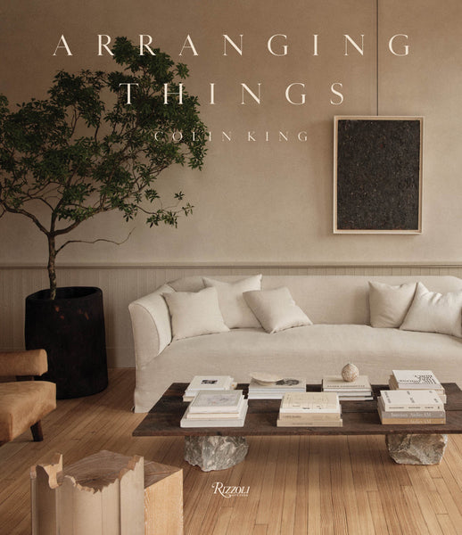 A minimalist living room showcasing personal creativity with a sofa, coffee table with books, wooden chair, large plant, and framed wall art. The cover text reads "Arranging Things" by Penguin Random House.