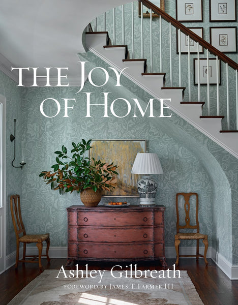 Book cover of "The Joy Of Home" by Common Ground, featuring an entryway with a red dresser, two chairs, a lamp, and a large potted plant. The elegant style is complemented by a staircase and framed artwork in the background, epitomizing Southern decorating charm.