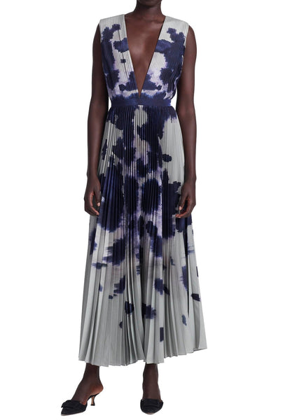 A person wearing an Altuzarra Pamela Dress, a sleeveless, deep V-neck, pleated midi dress with a dark blue and white abstract pattern, stands against a white background, paired with black heeled mules.