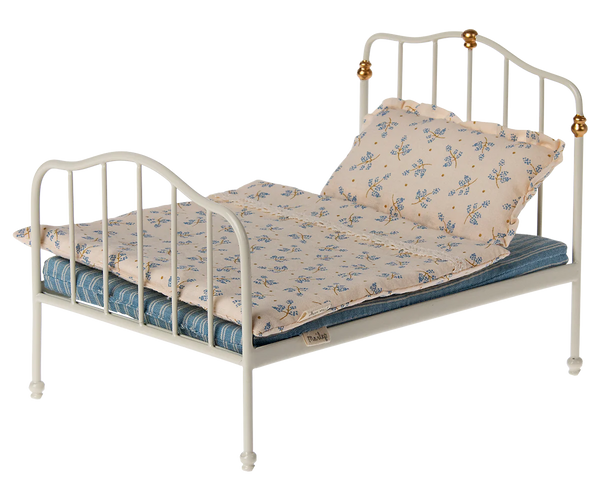 The Maileg Bed, Mouse - Off White is a small, white metal-framed bed adorned with blue and white floral bedlinen and a blue striped mattress made from Maileg's printed fabrics. It's perfectly sized for a child or their collection of mice and bunnies.