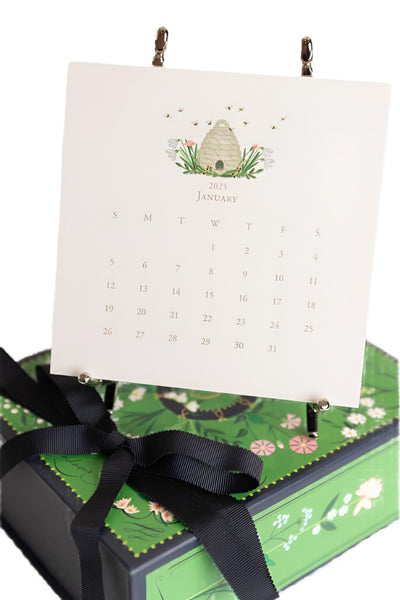 A 2025 Karen Adams calendar displaying the month of January stands on a decorative silver easel. The hand-finished calendar from the Karen Adams brand features an original design with an illustration of a beehive adorned with flowers and bees.