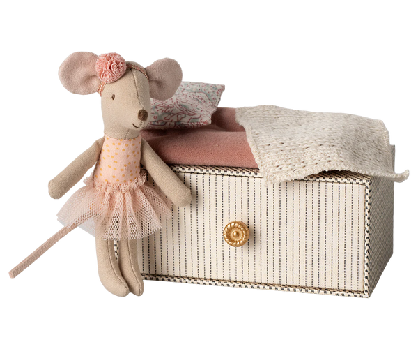 The Maileg Dance Mouse in Daybed, Little Sister, features an adorable little sister mouse dressed in a pink dress and flower headband, comfortably leaning against a small striped box with a gold knob. The box is covered by pink and white fabric, making it perfect for use as a day bed. This charming product from Maileg is recommended for children ages 3 and up.