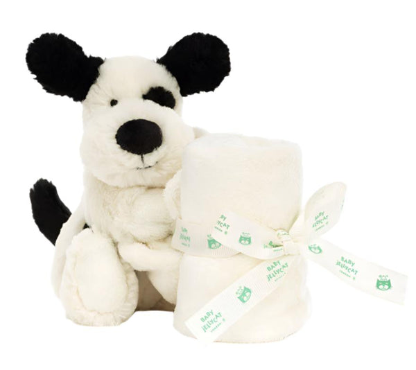 A Jellycat Bashful Black & Cream Puppy Soother sits next to a rolled-up white blanket tied with a ribbon that has "BABY BLANKET" printed on it, made from recycled fibers.