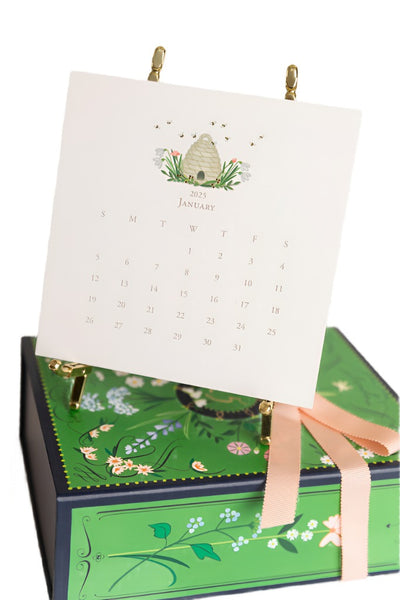 Displayed on a decorative green and blue box with a pink ribbon, the January 2025 page of the Karen Adams 2025 Classic Desk Calendar features a beehive illustration at the top and is elegantly presented on a gold easel. The hand-finished design adds an elegant touch to your desk décor.