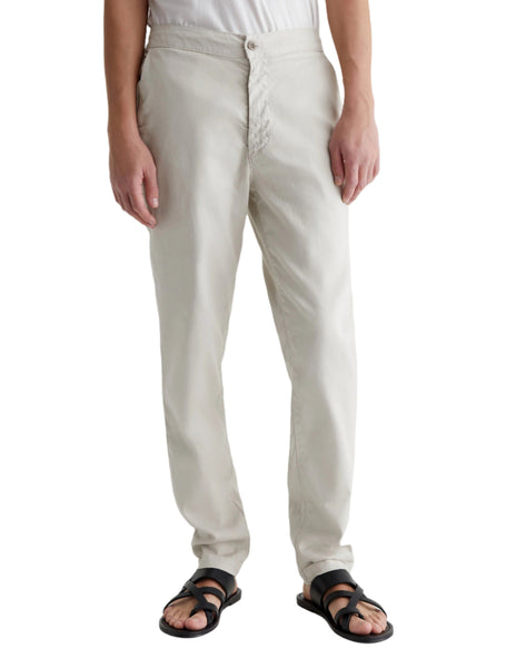 Person wearing light-colored, slim-cut AG Garreth Pant by AG Jeans with an adjustable drawstring, a white shirt, and black sandals, standing against a plain white background.