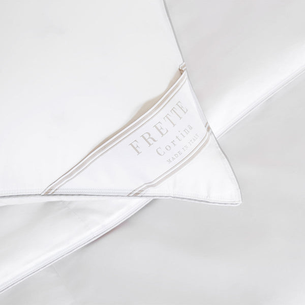 Close-up of a white Frette cotton sateen pillow with a label that reads "FRETTE Cortina Made in Italy." The Frette Cortina Pillow Insert, Firm Density is placed on a neatly arranged bed with white linens.