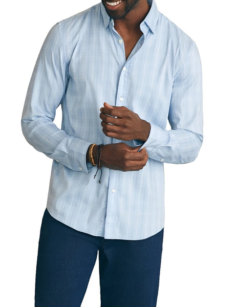 A person wearing a light blue Faherty Movement Sport Shirt and dark pants is adjusting the sleeve of their shirt with a slight smile.