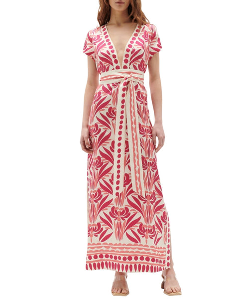 A woman is standing and wearing the Figue Isolda Dress, which features a floor-length design with a pink and white pattern, deep V-neckline, short sleeves, and a flowing column silhouette. She is also wearing flat sandals.