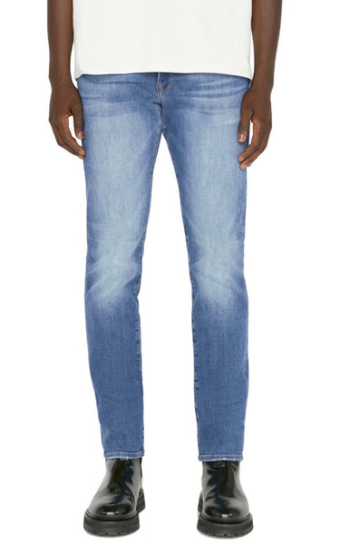 A person wearing Frame Men's L’Homme Slim Denim Capistrano jeans made from Comfort stretch denim and black boots, paired with a white top, is shown from the waist down against a white background.