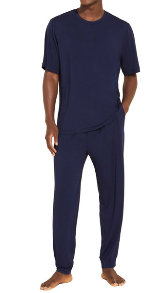 A person stands wearing the Eberjey Henry Long PJ Set from Eberjey, which includes a navy blue short-sleeve t-shirt and matching long pants made of a soft, breathable fabric with an elastic waistband for added comfort.