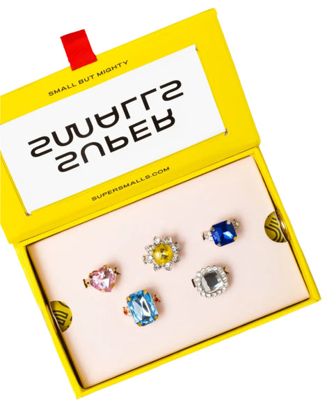 A yellow box labeled "SMALL BUT MIGHTY" showcases the Super Smalls Power Lunch Ring Set, featuring five sparkly rings adorned with colorful gemstones, set against a pink background. The website "SUPERSMALLS.COM" is prominently displayed on the box.