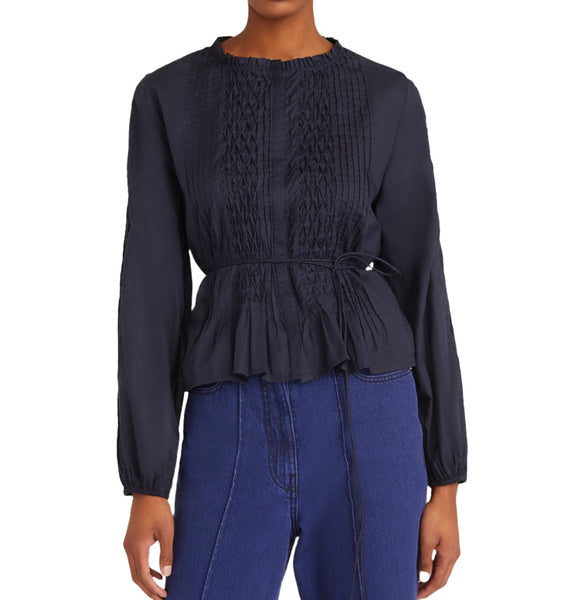 A person wearing the Merlette Bienne Top by Merlette New York, a black long-sleeve blouse inspired by Victorian-era designs with hand-stitched honeycomb smocking and a tied waist, paired with blue jeans.