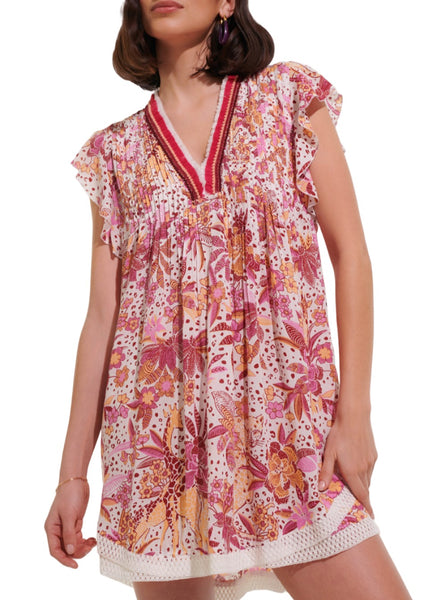 A person wearing the Poupette St Barth Sasha Mini Dress exudes true beach style—this short dress features a floral pattern with shades of pink, red, and orange, complemented by a V-neckline and ruffled sleeves. The creation from Poupette St Barth is adorned with intricate detailing around the neckline and hem.