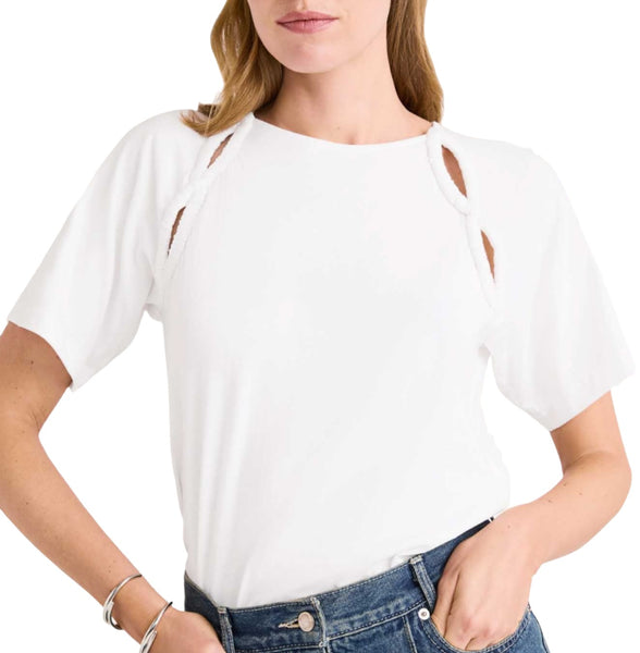 A woman wearing a white Merlette Solace Jersey Top from Merlette New York with shoulder cut-outs and ruched details, paired with blue jeans, stands with one hand on her hip. She has light-colored hair and is accessorized with bracelets on her right wrist.