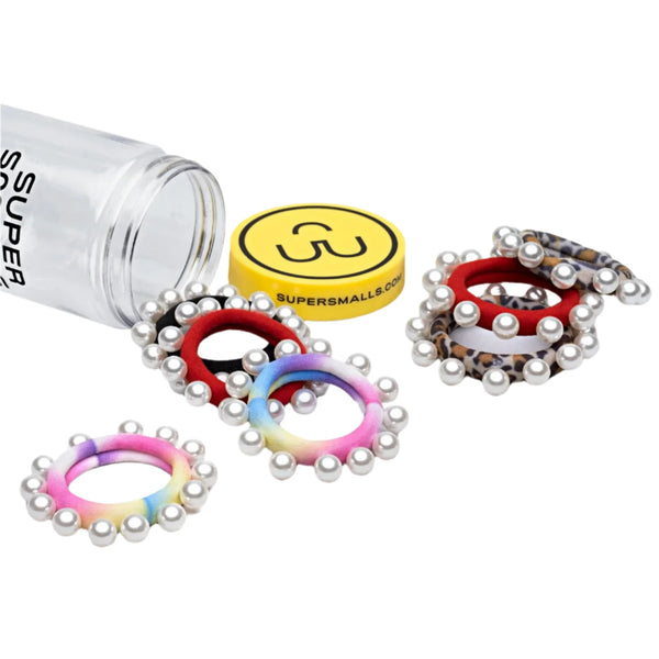 A clear container labeled "Super Smalls" lies on its side with colorful, fashion-forward hair ties adorned with pearl-like beads spilling out beside a yellow circular box lid also featuring the "Super Smalls" branding. These hair accessories, known as the "Super Smalls Central Park Pearl Hair Ties," blend mixed patterns effortlessly for a chic touch.