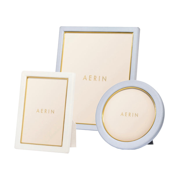 Three frames from the AERIN Classic Piero Leather Frame Collection by Aerin are elegantly displayed: a rectangular frame with a white border, a square frame with a blue border, and a round frame adorned with blue borders featuring gold detailing.