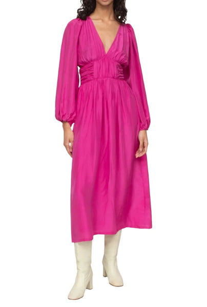 A person wearing the Sea NY Fabiola Dress, a long-sleeved pink silk habotai dress with a deep V-neckline, paired with white boots stands against a plain background.