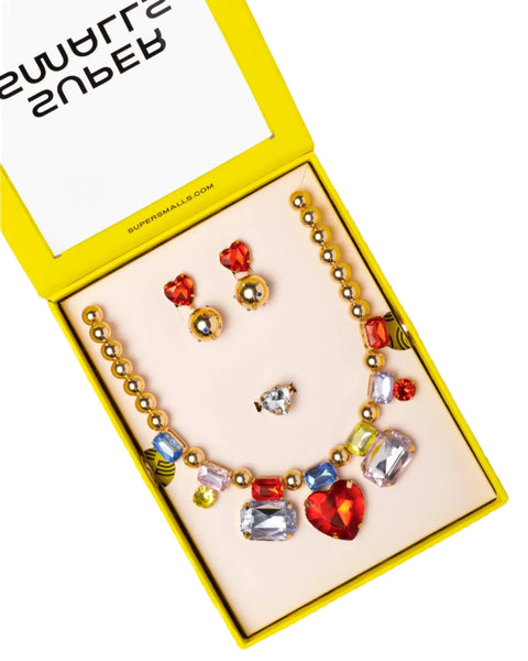 The Super Smalls Red Carpet Mega Set by Super Smalls comes in a yellow box, featuring a necklace with oversized gemstones, heart-shaped earrings, a ring, and ruby red stud earrings. The lid of the box displays text.