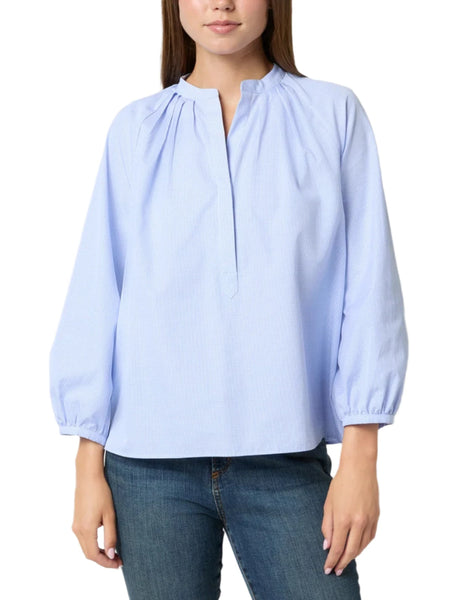 A person wearing the Ann Mashburn Sienna Top in light blue, made of 100% cotton with a pleated split neckline, paired with blue jeans, and standing against a white background.