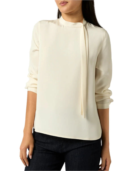 A woman wearing the Ann Mashburn Pippa Top, a 100% silk, cream-colored blouse featuring an asymmetrical tie detail at the neck, paired with black pants.