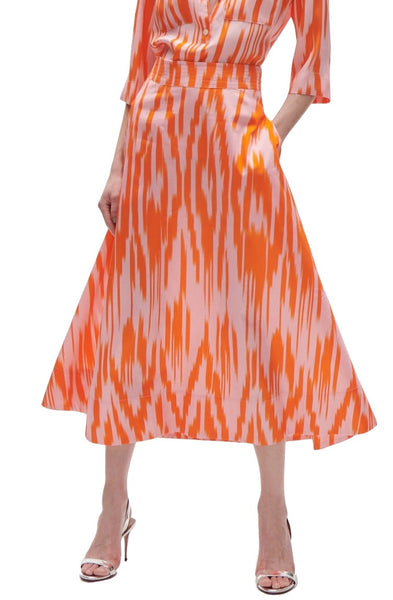 A person stands wearing a flowy, mid-length Figue Isla Skirt with an orange and white geometric pattern, featuring a trapunto waistband. The A-line silhouette is paired with a matching top and silver high-heeled shoes, creating an effortlessly stylish look.