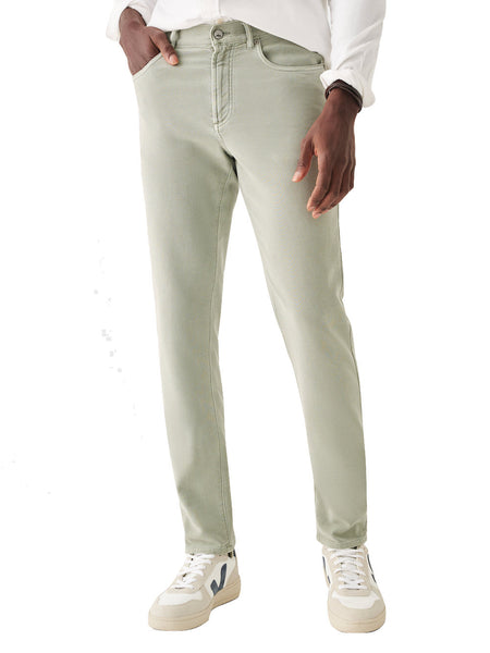 A person is wearing light green, bestselling Faherty Stretch Terry 5 Pocket Pants, a white shirt, and white sneakers with black details. The person's left hand is in the left pocket, and they are standing against a white background.