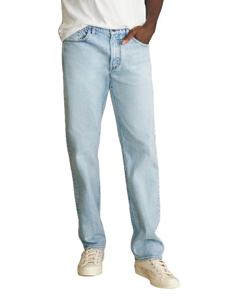 Person wearing Faherty Slim Straight Denim jeans in light blue, made from organic cotton, and white sneakers, with one hand in a pocket, isolated on a white background.