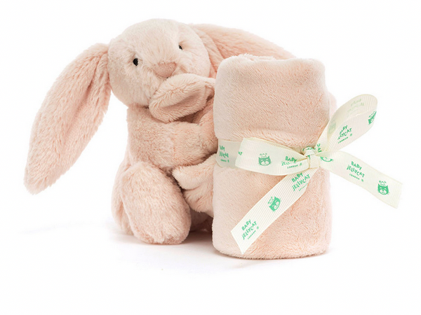 A Jellycat Bashful Blush Bunny Soother cuddles a rolled, beige baby blanket, tied with a white ribbon featuring green text and owl illustrations, making it the perfect newborn gift.